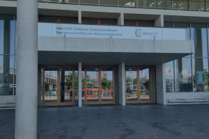 Das Bild zeigt den Eingang des Konzrthauses in Freiburg mit einem Hinweise auf die Konferenz im Fenster über den Türen. Als Titelbild hat ein blaues Overlay, damit man die Schrift darauf erkennen kann.