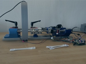 Eine Nahaufnahme zeigt eine Versuchsaufbau-Zugmaschine auf einer Werkbank, die mit blauen und roten 3D-gedruckten Komponenten, verschiedenen MEchanik- sowie Elektronikbauteilen und einer darüber montierten Kamera in gedruckter Halterung ausgestattet ist; davor liegen mehrere weiße, handbreite Kunststoffproben mit verbreiterten Enden zum Einspannen. Dieses Bild hat zusätzlich ein dunkelblaues und transparentes Overlay, um die Schrift darauf besser lesbar zu machen.
