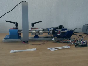 Eine Nahaufnahme zeigt eine Versuchsaufbau-Zugmaschine auf einer Werkbank, die mit blauen und roten 3D-gedruckten Komponenten, verschiedenen MEchanik- sowie Elektronikbauteilen und einer darüber montierten Kamera in gedruckter Halterung ausgestattet ist; davor liegen mehrere weiße, handbreite Kunststoffproben mit verbreiterten Enden zum Einspannen.
